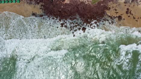 Aerial-drone-birds-eye-shot-of-small-waves-crashing-into-rocks-on-the-famous-tropical-Love-beach-in-Pipa-Brazil-in-Rio-Grande-do-Norte-with-golden-sand,-warm-turquoise-water-and-colorful-umbrellas