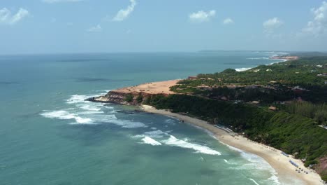 Left-trucking-aerial-drone-shot-of-the-famous-golden-Pipa-Plateau-with-tropical-beaches-below-looming-cliffs-covered-in-green-foliage-and-small-waves-crashing-ashore-in-Rio-Grande-do-Norte,-Brazil