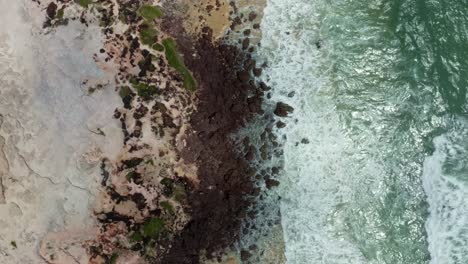 Aerial-drone-birds-eye-top-view-of-small-waves-crashing-into-rocks-on-the-famous-tropical-Love-beach-in-Pipa-Brazil-in-Rio-Grande-do-Norte-with-golden-sand,-brown-rocks,-and-warm-turquoise-water