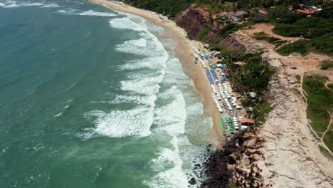 Dolly-in-tilt-down-aerial-drone-birds-eye-top-view-shot-approaching-the-tropical-famous-Love-Beach-in-Pipa,-Brazil-Rio-Grande-do-Norte-during-high-tide-with-waves-crashing-and-colorful-umbrellas