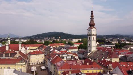Clock-tower-of-Klagenfurt-chapel-standing-out-above-red-roofs,-aerial-pullback