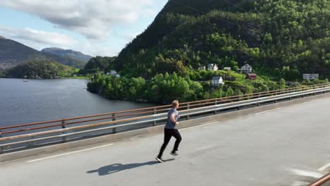 Drone-flies-past-man-jogging-over-Norway-fjord-bridge,-parallax,-to-front-view