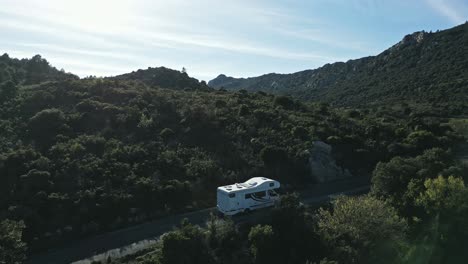 White-RV-Camping-Car,-Camper-Van-Drives-Uphill-Green-Mountains,-Valley-Landscape-in-Pyrenees-Summer,-Aerial-Drone-Establishing-Shot