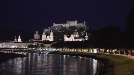 An-extremely-long-view-of-Fortress-Hohensalzburg,-Salzburg,-Austria-at-night-when-the-artificial-lighting-is-falling-into-the-castle-and-a-lake's-water