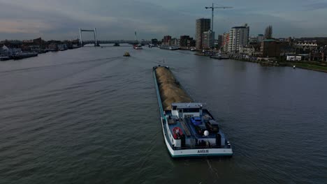 Aerial-view-of-sand-loaded-general-cargo-ship-Avelie-sailing-through-river-with-waterfront-skyline-buildings-and-city-view-in-the-background
