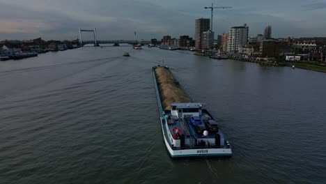 Luftaufnahme-Des-Mit-Sand-Beladenen-Stückgutschiffs-Avelie,-Das-Durch-Den-Fluss-Segelt,-Mit-Skyline-Gebäuden-Am-Wasser-Und-Blick-Auf-Die-Stadt-Im-Hintergrund