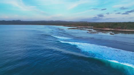Establishing-drone-shot-of-ocean-waves-and-coral-reef,-with-shoreline-of-the-national-park-at-the-tip-of-Java-island,-Indonesia
