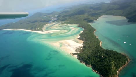 Handheld-clip-from-airplane-of-tropical-island-surrounded-by-turquoise-sea,-with-white-sand-beach-and-thick-forest