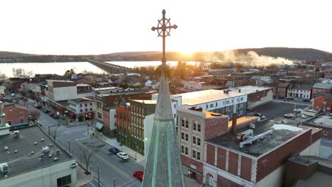 Panoramic-drone-view-of-a-city-in-America-at-sunset-in-the-spring,-with-a-focal-point-of-a-church-steeple-and-reflections-off-of-the-buildings-with-a-river-and-bridges-in-the-background