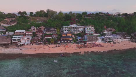 Panning-drone-clip-of-Bingin-beach,-Bali-with-lines-of-sun-loungers-and-popular-tourist-hotels,-then-moving-to-stunning-limestone-cliffs-and-turquoise-water-with-coral-reefs