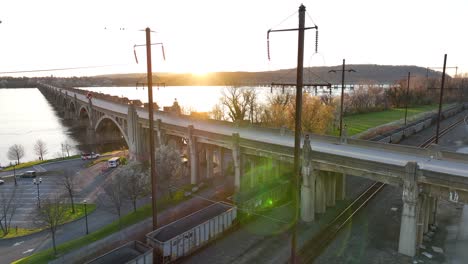 Aerial-view-of-two-bridges-crossing-a-scenic-river-and-the-riverfront-with-green-space-at-sunset-in-the-spring,-with-railways-and-freight-trains