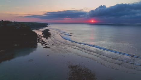 Calming-drone-clip-showing-quiet-beach-in-Bali,-Indonesia,-with-beautiful-pink-sunset-over-calm-ocean