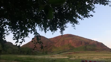 mesmerizing-Arthur's-Seat-Hill-in-Holyrood-Park,-showcasing-the-stunning-landscape-through-the-gentle-sway-of-tree-branches-and-leaves,-bathed-in-the-warm,-enchanting-light-of-a-breathtaking-sunset
