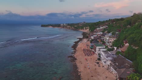 Overhead-drone-clip-of-popular-tourist-destination,-Bingin-beach-in-Bali,-showing-hotel-and-tourism-infrastructure-next-to-coral-reef-and-perfect-surf-waves