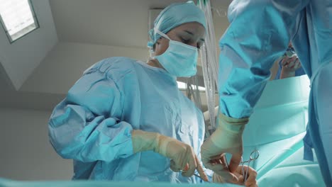 surgical-intervention-low-angle-view-of-a-female-surgeon-with-all-the-blue-protective-equipment,-mask,-gown,-cap,-gloves,-who-uses-scissors-with-the-help-of-another-person-from-behind
