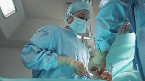 Intervención-Quirúrgica-Vista-De-ángulo-Bajo-De-Una-Cirujana-Con-Todo-El-Equipo-De-Protección-Azul,-Máscara,-Bata,-Gorra,-Guantes,-Que-Usa-Tijeras-Con-La-Ayuda-De-Otra-Persona-Desde-Atrás