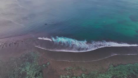 Overhead-wide-angle-drone-shot-of-local-Indonesian-fishing-boat-cruising-the-coastline-and-coral-reef,-with-surfers-paddling-and-waves-rolling-towards-shore-in-Bali