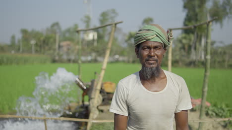 Pobre-Granjero-Bangladesí-Mirando-La-Cámara-Mientras-La-Bomba-De-Agua-Funciona-En-El-Fondo-Arrojando-Agua-Al-Campo-De-Cultivo
