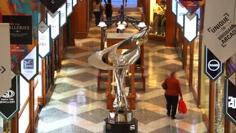 Dynamic-zoom-in-shot-capturing-Mirage-sculpture-by-Gidon-Graetz-as-the-centrepiece-of-Brisbane-arcade,-shoppers-walking-across-fashion-laneway,-window-shopping-and-browsing-at-the-designer-boutiques