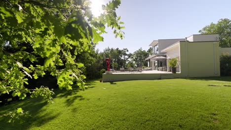Beautiful-green-lawn-with-a-luxurious-white-villa-seen-from-the-side,-rectangular-shape,-with-large-terrace-and-sun-loungers,-blue-sky-and-bright-sun