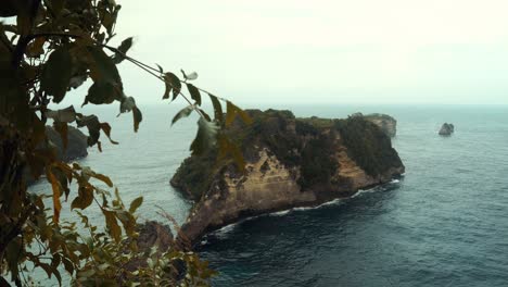 Zeitlupenaufnahme-Einer-Insel-Auf-Nusa-Penida-Aus-Bali-In-Indonesien-Mit-Blick-Auf-Eine-Andere-Insel-Mit-Klippen-Und-Bäumen-Darauf-Bei-Ruhigen-Wellen-An-Einem-Windigen-Morgentag