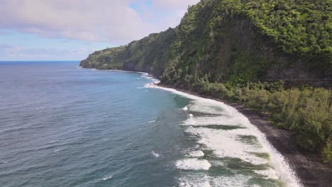Drone-capture-revealing-the-scenic-side-of-Waipi'o-Valley-from-a-black-sand-beach-under-partial-sunlight,-with-moderate-waves-on-Hawaii's-Big-Island