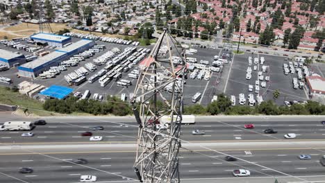 Drone-descends-towards-a-power-line-tower,-revealing-a-blend-of-highway,-commercial,-and-residential-scenery-in-the-background