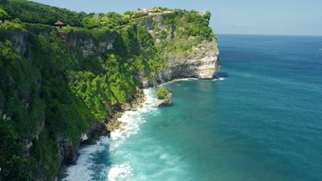 Tilt-shot-at-Pura-Uluwatu-temple-from-the-cliffs-in-Bali-overlooking-the-blue-sea-with-waves-and-the-beautiful-island-on-a-sunny-day