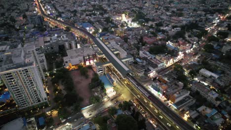 The-city's-metro-train-stops-at-the-metro-rail-station,-traffic-streets-and-apartment-buildings,-as-seen-in-this-evening-aerial-view-of-Chennai-City's-busy-Vadapalani-Signal-neighborhood
