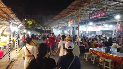 Indonesian-People-Walk-at-Street-Food-Night-Market,-Denpasar-Bali-Eating-Snacks