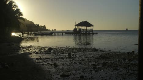Punta-Gorda-Beach-in-Roatan,-Honduras-during-sunset-with-wooden-pier-and-rustic-gazebo-made-out-of-palm-leaves-and-commercial-boat-afar---Golden-hour-silhouette-wide-shot