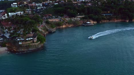 Left-trucking-aerial-drone-shot-following-a-boat-sailing-on-the-Guaraíras-Lagoon-towards-the-famous-sunset-lookout-in-the-tropical-beach-town-of-Tibau-do-Sul-in-Rio-Grande-do-Norte,-Brazil-during-dusk