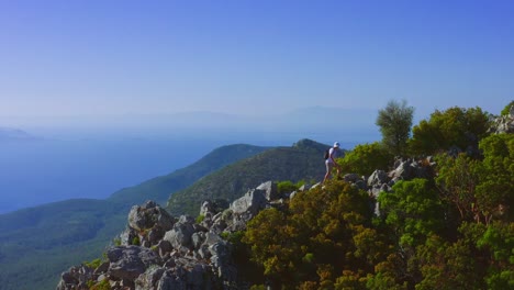 Aerial-view-of-man-walking-to-the-mountain-peak-against-scenic-Aegean-landscape,-Carian-trail,-Datça