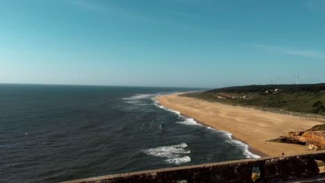Aerial-wide-shot-of-sandy-beach-in-Nazare-with-waves-of-Atlantic-Ocean-during-sunset-time---Rotating-wind-turbine-farm-in-background