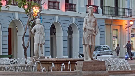 Straßenansicht-Des-Plaza-De-Armas-Brunnens-Mit-Statuen-Als-Fußgänger,-Autos-Und-Einem-Motorroller-Im-Hintergrund