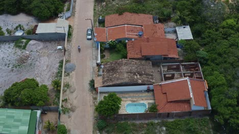 Tilt-down-aerial-drone-birds-eye-top-view-shot-following-a-motorcycle-driving-down-a-dirt-road-in-the-rural-residential-area-of-the-tropical-beach-town-of-Tibau-do-Sul-in-Rio-Grande-do-Norte,-Brazil
