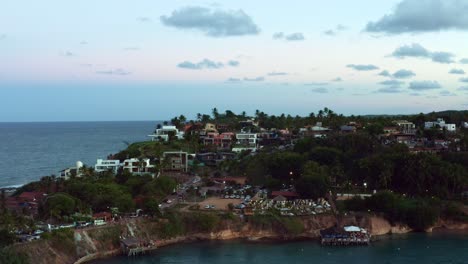 Left-trucking-aerial-drone-shot-of-the-famous-tropical-beach-town-of-Tibau-do-Sul-in-Rio-Grande-do-Norte,-Brazil-with-hills-of-large-homes-and-hotels-during-a-warm-summer-evening