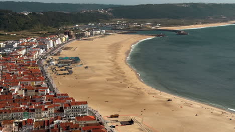 Aerial-wide-shot-of-flying-seagulls-over-sandy-beach-and-old-Portuguese-city-at-sunset---Nazare,-Portugal---tilt-up