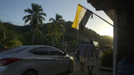 Die-Garifuna-Flagge-Hängt-Am-Nachmittag-Gehisst-Vor-Einem-Haus-In-Punta-Gorda,-Roatán,-Honduras-–-Von-Hinten-Beleuchtet