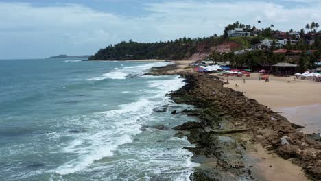 Tilt-up-aerial-drone-shot-of-the-tropical-Tibau-do-Sul-peninsula-with-waves-crashing-into-algae-covered-rocks,-tourists-enjoying-shade-under-colorful-umbrellas-in-Rio-Grande-do-Norte,-Brazil