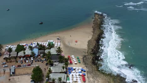 Tilt-down-aerial-drone-birds-eye-top-view-of-small-waves-crashing-into-algae-rocks-from-the-tropical-Tibau-do-Sul-beach-near-Pipa,-Brazil-in-Rio-Grande-do-Norte-with-colorful-umbrellas-for-tourists