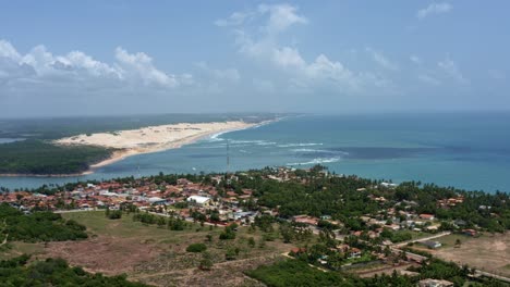 Lowering-aerial-drone-extreme-wide-shot-of-the-tropical-beach-town-of-Tibau-do-Sul-in-Rio-Grande-do-Norte,-Brazil-with-the-Malembá-Sand-Dunes,-Atlantic-Ocean,-and-Guaraíras-Lagoon-in-the-background