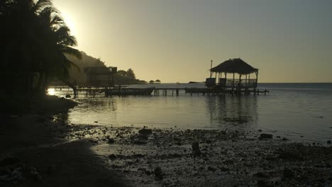 Punta-Gorda-Beach-in-Roatan,-Honduras-during-sunset-with-wooden-pier-and-rustic-gazebo-made-out-of-palm-leaves---Golden-hour-silhouette
