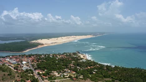 Rotating-aerial-drone-extreme-wide-shot-of-the-tropical-beach-town-of-Tibau-do-Sul-in-Rio-Grande-do-Norte,-Brazil-with-the-Malembá-Sand-Dunes,-Atlantic-Ocean,-and-Guaraíras-Lagoon-in-the-background