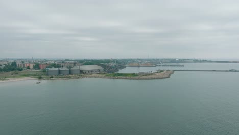 Aerial-establishing-view-of-port-cranes-and-empty-loading-docks-at-Port-Of-Liepaja-,-Liepaja-city-in-the-background,-overcast-summer-day,-wide-drone-shot-moving-forward