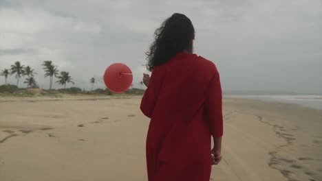 A-brunette-with-long-hair,-dressed-in-a-red-dress,-walks-on-an-empty-beach,-holding-a-balloon-on-a-string,-while-a-gentle-breeze-blows-and-tousles-her-hair