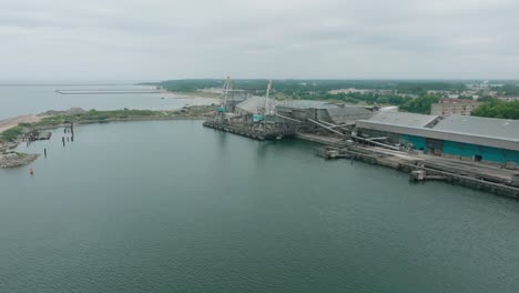 Aerial-establishing-view-of-port-cranes-and-empty-loading-docks-at-Port-Of-Liepaja-,-Liepaja-city-in-the-background,-overcast-summer-day,-wide-drone-shot-moving-forward,-tilt-down