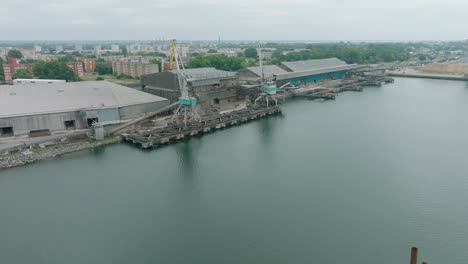 Aerial-establishing-view-of-port-cranes-and-empty-loading-docks-at-Port-Of-Liepaja-,-Liepaja-city-in-the-background,-overcast-summer-day,-wide-slow-ascending-drone-shot-moving-backward