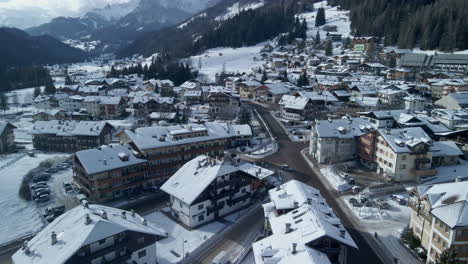 Aerial-view-of-drone-flying-over-Winter-cityscape-in-Val-di-Fassa,-Italy,-with-snow-covered-buildings-and-Dolomites-backdrop