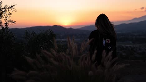 girl-sitting-on-a-cliff-edge-at-Mt-Rubidoux-in-Riverside-California-with-a-black-Fjallraven-backpack-during-a-bright-orange-and-pink-sunset-with-foxtail-grass-in-the-foreground-blowing-in-the-wind-4k